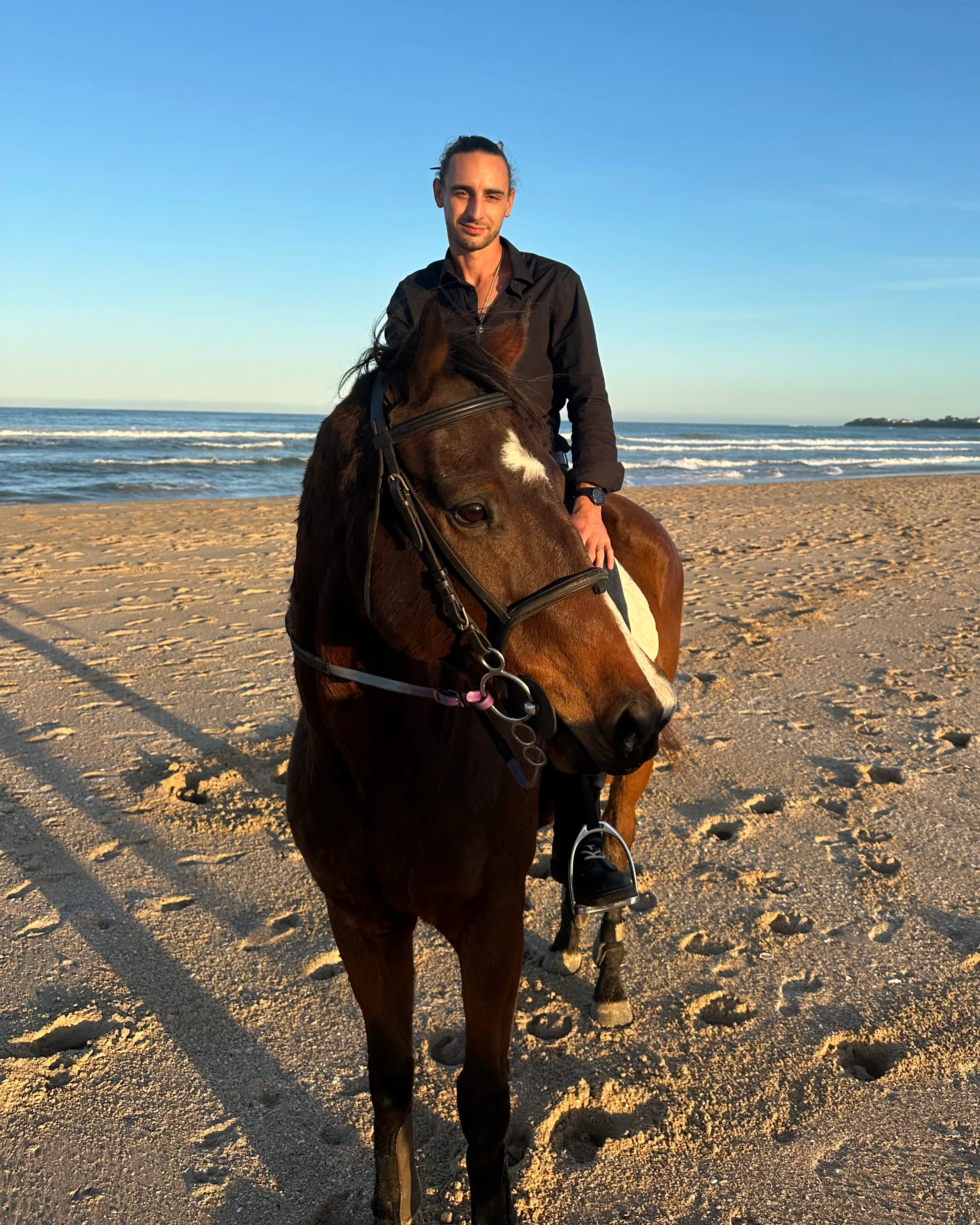 Theuns Pienaar riding a dark bay horse on a beach, smiling at the camera